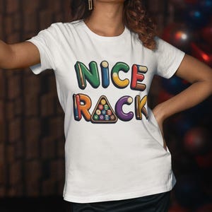 Puede incluir: Camiseta blanca con el texto "NICE RACK" en letras coloridas y con forma de burbujas. La palabra "RACK" presenta un gráfico de un triángulo de billar con bolas. La persona lleva la camiseta y sostiene un taco de billar cerca de una mesa verde.