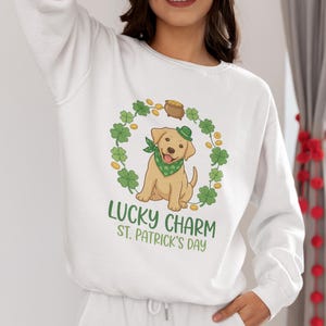 Könnte beinhalten: Weißes Sweatshirt mit einem St. Patrick's Day-Motiv. Das Design zeigt einen Cartoon-Labrador-Welpen mit grünem Halstuch und Hut, umgeben von Kleeblättern und Goldmünzen. Der Text lautet "Lucky Charm St. Patrick's Day".