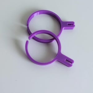 Può includere: Due oggetti circolari viola con un'estensione a forma di manico. Gli oggetti sono fatti di un materiale solido e hanno una superficie liscia. Un oggetto è completo, mentre l'altro ha un'apertura nel cerchio.