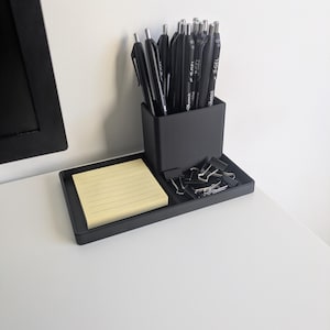 May include: A black desk organizer with a pen holder, a small tray with a notepad, and binder clips. The pen holder is filled with black pens. The notepad is yellow with horizontal lines. The organizer sits on a white surface.