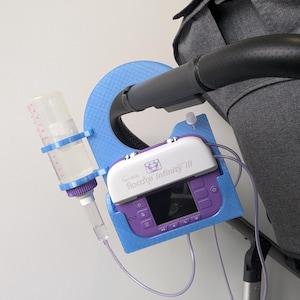 May include: A blue 3D-printed mount attached to a pushchair handle, holding a Nutricia Flocare Infinity III enteral feeding pump and a clear plastic bottle with measurement markings, connected by purple tubing for medical nutrition delivery.