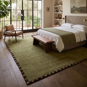 May include: A bedroom with a large window overlooking a garden. The room features a green rug with a dark border, a bed with white linens and a green throw, a wooden bench, and a leather chair. A floor lamp and artwork complete the space.