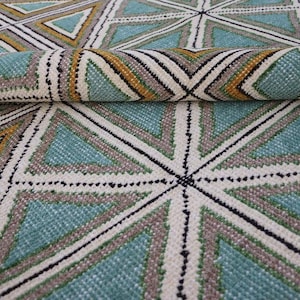 May include: A rolled-up rug with a geometric pattern. The design features teal, white, gray, and gold triangles and lines. The rug's texture is visible, suggesting a woven construction. The overall aesthetic is modern and decorative.