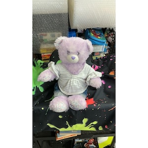 Può includere: Un orsacchiotto di peluche con pelliccia viola chiaro, che indossa una maglietta bianca a maniche corte con sottili righe grigie orizzontali e vita arricciata. L'orso ha un naso viola e le zampe presentano un motivo multicolore a coriandoli.