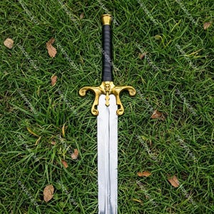 May include: A decorative sword with a gold-colored hilt featuring a skull design and a black leather-wrapped handle. The blade is silver and the sword is set against a backdrop of green grass.