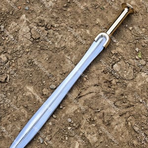 May include: A silver sword with a gold handle and guard. The double-edged blade tapers to a point. The handle has a rounded pommel. The sword is displayed on a dirt surface.