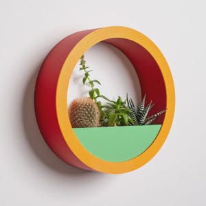 May include: A round, wall-mounted planter with a red exterior and orange trim. The interior features a green base holding various succulents and a small cactus. The planter is approximately 12 inches in diameter.