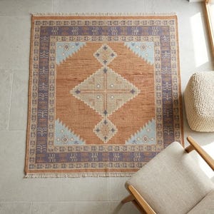 Könnte beinhalten: Ein rechteckiger Teppich mit geometrischem Design. Der Teppich hat ein zentrales Rautenmuster in Beige und Hellbraun, umgeben von einem Rand aus lila und hellblauen geometrischen Formen. Der Teppich hat einen Fransenrand und liegt auf einem gefliesten Boden.