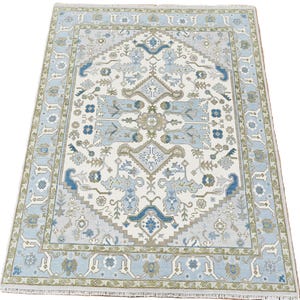 Könnte beinhalten: Ein rechteckiger Teppich mit einem aufwendigen, symmetrischen Design. Der Teppich hat ein elfenbeinfarbenes Mittelfeld mit einem sich wiederholenden Muster in Blau-, Grau- und Olivgrüntönen. Ein hellblauer Rand rahmt das Design ein und sorgt für ein stimmiges Aussehen. Der Teppich hat einen Fransen.