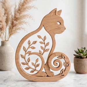 Puede incluir: Una figura de gato de madera de color marrón claro con un diseño recortado de un gato sentado de perfil. El interior de la forma del gato contiene ramas intrincadas cortadas con láser con pequeñas hojas que se curvan en la cola y el cuerpo.
