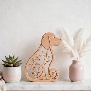 Könnte beinhalten: Eine hellbraune Holzfigur eines sitzenden Hundes mit einem eingearbeiteten Ranken- und Blattmuster im Körper. Die Hundefigur steht zwischen einer kleinen Sukkulente in einem zweifarbigen Topf und einer hellrosa Vase mit weißem Trockengras.