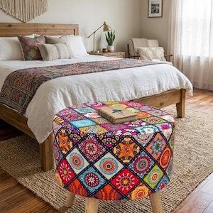Puede incluir: Un otomán colorido y estampado con patas de madera en un dormitorio. La parte superior del otomán presenta un diseño de patchwork con cuadrados de varios colores y patrones. Un libro descansa encima. La habitación tiene una cama y un tapiz tejido.