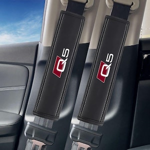 以下が含まれることがあります： 白ステッチと赤と白の「Q5」ロゴが入った、2つの黒いシートベルトショルダーパッド。パッドは車内に取り付けられており、シートベルトのバックルが下部に表示されています。背景には車の内装が写っています。