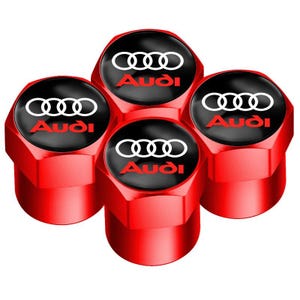 May include: Four red valve stem caps with the Audi logo. Each cap has a black top with the Audi logo in white and red lettering. The caps are made of metal and have a hexagonal shape.