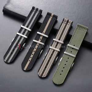 May include: Four nylon watch straps with silver-toned metal buckles and keepers. The first is black with a central gray stripe, the second is black with a tan stripe, the third is black with a tan and gray stripe, and the fourth is solid olive green.