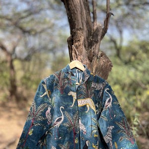 Puede incluir: Chaqueta color turquesa con un estampado de jungla vibrante con leopardos, cebras, pájaros y follaje tropical. La chaqueta está colgada en una percha de madera.
