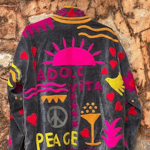 Puede incluir: Chaqueta gris oscuro con diseños bordados coloridos. La parte trasera presenta un sol rosa, las palabras "DOLCE VITA", un símbolo de la paz, la palabra "PEACE", corazones y un pájaro amarillo. La chaqueta tiene un ribete amarillo y rosa.