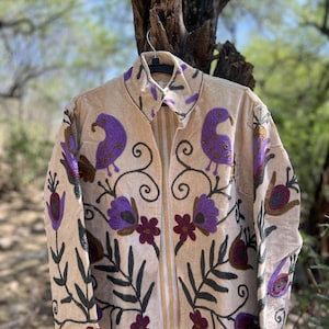 May include: Beige jacket with purple bird and floral embroidery. The jacket features a collared neckline and is adorned with intricate embroidered designs in shades of purple, maroon, and green. The jacket is hanging on a black hanger.