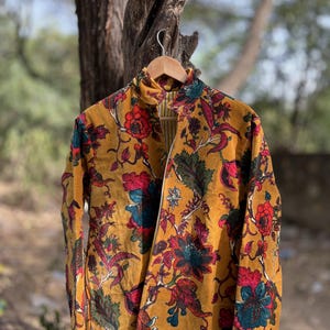 Könnte beinhalten: Senffarbene Jacke mit einem lebendigen Blumenmuster in Rot, Türkis und Grün. Die Jacke hat einen hohen Kragen und lange Ärmel. Der Stoff scheint Samt oder ein ähnliches weiches Material zu sein, mit einem goldenen Reißverschluss.