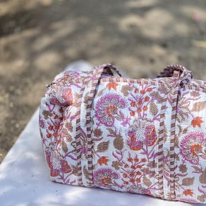 May include: A white duffel bag featuring a floral design in pink, orange, and brown. The bag has brown trim and handles. The floral pattern is a repeating design of stylized flowers and leaves.