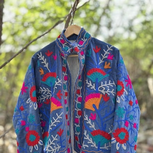 Pode incluir: Uma jaqueta azul bordada com desenhos florais coloridos. A jaqueta apresenta flores vermelhas, laranja, rosa e turquesa com detalhes brancos. O forro interno tem um padrão listrado. A jaqueta está pendurada em um cabide de madeira.