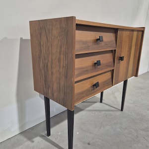 Può includere: Un mobile in legno moderno della metà del secolo con tre cassetti e una porta laterale. Il mobile ha una finitura in legno scuro, maniglie quadrate nere e gambe affusolate nere. Il mobile misura circa 76 cm di larghezza, 38 cm di profondità e 76 cm di altezza.