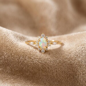 Könnte beinhalten: Ein goldener Ring mit einem zentralen, ovalen, schillernden Opal, der von einem Kranz aus kleinen runden, klaren Steinen umgeben ist, ergänzt durch zwei marquise-geschliffene klare Steine an den Seiten, die ein sternförmiges Design bilden.