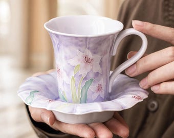 May include: A porcelain teacup and saucer set featuring a soft lavender watercolor-style background with hand-painted white lily flowers and green stems. The cup has a scalloped rim and a curved handle, while the saucer has a matching wavy edge.