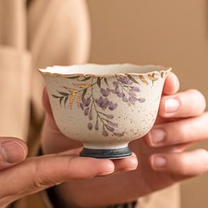 Puede incluir: Una pequeña taza de té de cerámica blanquecina con un acabado moteado. La taza presenta un delicado diseño floral en morado y dorado, con un borde festoneado y una base azul oscuro. La taza de té se sostiene con dos manos.