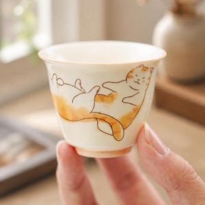 May include: A small, off-white ceramic tea cup featuring a hand-painted illustration of an orange and white tabby cat lying on its back. The cat has a smiling expression, and the cup has a smooth, rounded rim with a subtle orange-tinted base.