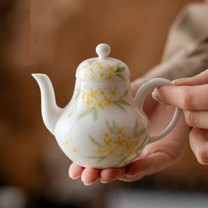 May include: A small, white ceramic teapot with a rounded body, curved spout, and handle. The teapot is decorated with yellow floral and green leaf patterns. The lid has a small, round knob. The teapot is held in a person's hand.