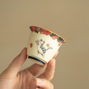 Peut inclure: Une petite tasse à thé en céramique blanc cassé avec une illustration de chat ludique. La tasse présente un bord bleu à la base et un motif floral rouge, noir et orange autour du bord. Un chat attrape des papillons.