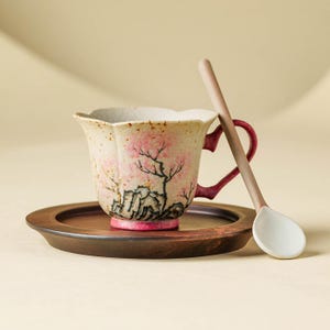 May include: A ceramic teacup with a floral design, a matching saucer, and a wooden spoon. The cup features a pink and black tree design on a cream-colored background. The handle is a deep red color. The saucer is dark brown.