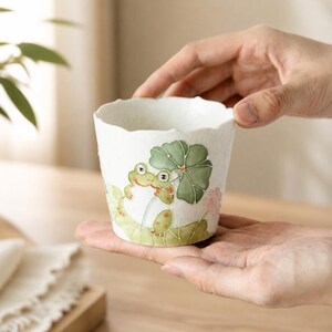 May include: A white ceramic cup with a scalloped rim, featuring a hand-painted illustration of a green frog holding a large green lily pad as an umbrella, surrounded by light green lily pads and small pink floral accents on the exterior surface.
