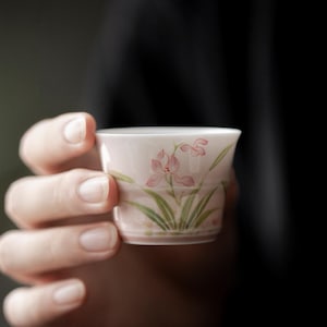 May include: A small, pale pink teacup with a floral design. The cup features a delicate illustration of pink orchids with green leaves. The cup is held in a person's hand, with a dark background.