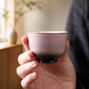 May include: A small, round ceramic tea cup featuring a textured, speckled finish that transitions from a soft, dusty mauve pink at the rim to a deep, dark charcoal brown at the base, with a smooth, light-coloured interior rim.