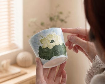 May include: A ceramic cup featuring a hand-painted light blue background with a prominent yellow lotus flower and large dark green leaves. The cup has a scalloped rim and a smooth white interior, showcasing detailed floral artwork on the exterior.