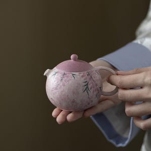 May include: A small, round, pink teapot with a matching lid. The teapot is decorated with a floral pattern in shades of pink and green. It has a small spout and a handle. The teapot is held in someone's hands.