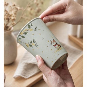 May include: A light sage green ceramic tumbler featuring a delicate illustration of a calico cat wearing a red collar, surrounded by scattered yellow floral branches and falling petals. The cylindrical vessel has a smooth finish and a tapered base.