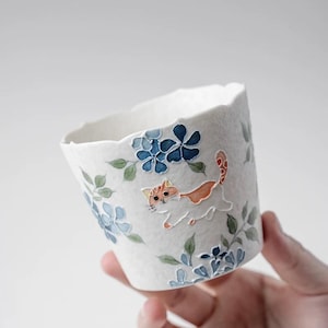 May include: White ceramic cup with a scalloped rim, decorated with blue floral patterns and a playful orange and white cat illustration. The cup is designed for beverages and has a textured surface.