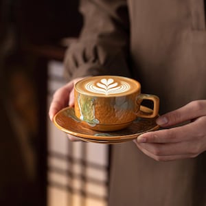 Könnte beinhalten: Ein Keramik-Kaffeebecher und Untertassen-Set mit Latte. Der Becher und die Untertasse sind eine Mischung aus Orange-, Braun- und Grüntönen. Die Latte Art zeigt ein Herzdesign. Das Set wird in den Händen einer Person gehalten.