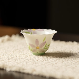 Peut inclure: Une tasse à thé en céramique blanche avec un bord festonné. La tasse présente un motif de fleur rose peint à la main avec des accents jaunes et verts. La tasse est posée sur une surface texturée de couleur crème.