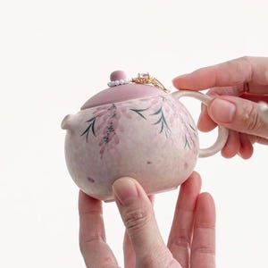 May include: A small, round, pink ceramic teapot with a matching lid. The teapot is decorated with a floral design in shades of pink and green. A gold ring with a pink gemstone sits on the lid. The teapot is held by two hands.
