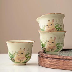 May include: A set of four ceramic teacups with a light green glaze and a scalloped rim. Each cup features a hand-painted illustration of a cute bunny surrounded by green leaves and white flowers. The cups are displayed on a wooden tray.