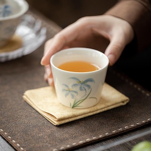 May include: A white ceramic teacup with a blue floral design, filled with tea. The cup sits on a square, tan coaster. A person's hand holds the cup. The tea has a golden colour.