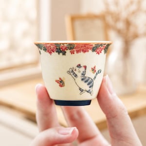 May include: A small, cream-coloured ceramic tea cup featuring a hand-painted illustration of a grey tabby cat reaching for a red butterfly. The rim is decorated with a band of red and orange floral patterns, and the base is a solid dark blue.