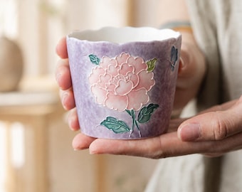 May include: A ceramic cup with a scalloped rim featuring a light purple mottled background and a central raised relief design of a large pink peony flower with green leaves and a small blue butterfly accent on the side of the vessel.