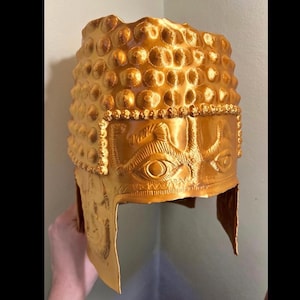 Op de afbeelding: Een goudkleurige helm met een met noppen bezette kroon en een gedetailleerde frontplaat. De helm heeft een oogontwerp en ingewikkelde patronen, wat wijst op een historisch of fantasiethema. De helm wordt door een hand vastgehouden.