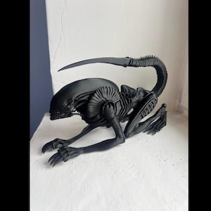 Puede incluir: Una figura negra de Xenomorfo, posada sobre una superficie blanca. La escultura detallada presenta una cola larga y curvada, un cuerpo acanalado y garras afiladas. El alienígena está en posición de gateo, mostrando su diseño amenazante.