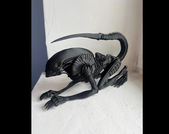 May include: A black Xenomorph creature figurine, posed on a white surface. The detailed sculpture features a long, curved tail, a ribbed body, and sharp claws. The alien is in a crawling position, showcasing its menacing design.